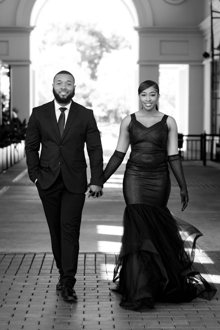 Black and white portrait of a couple in formalwear walking hand in hand